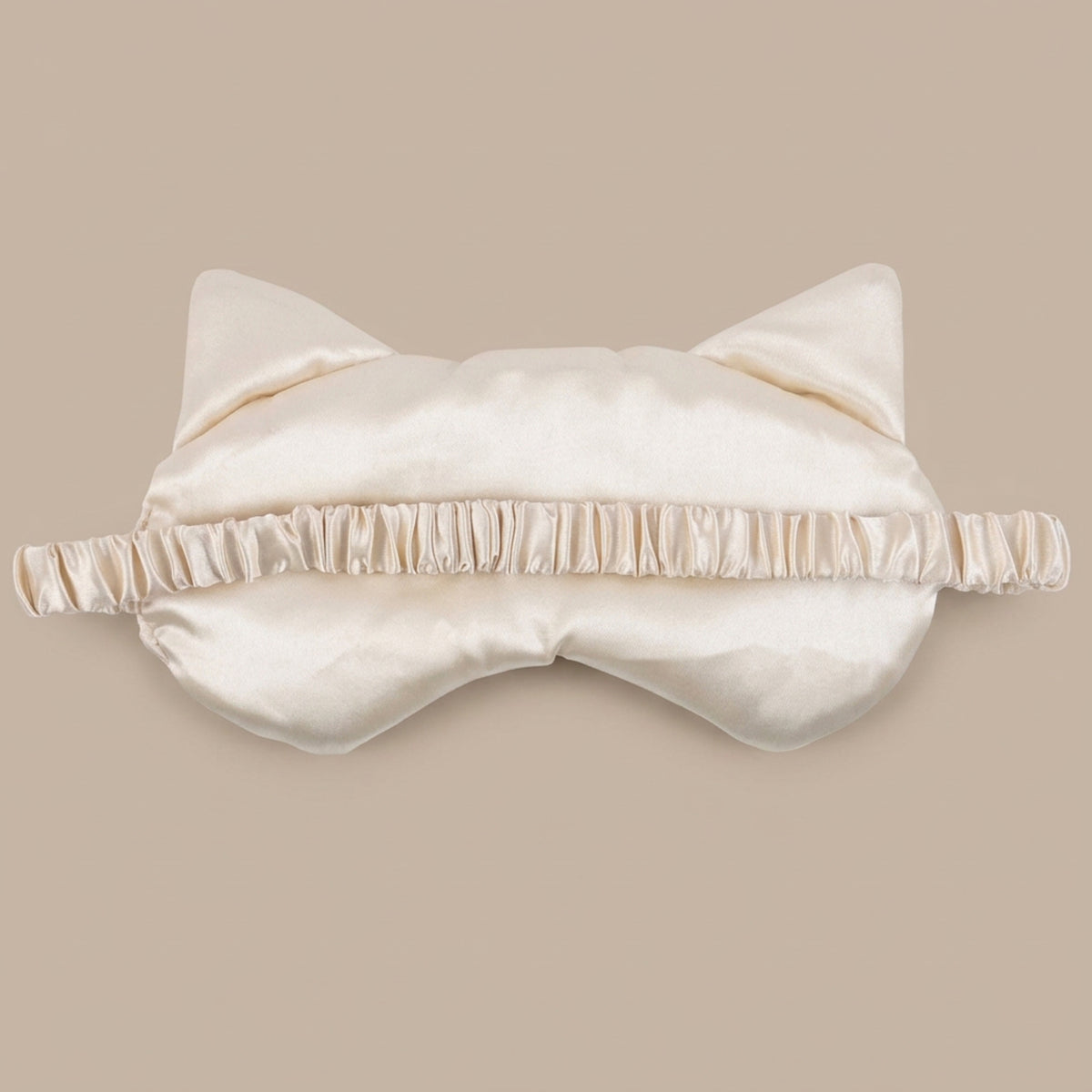 a back of an ivory satin sleep mask with cat ears and a ruffled elastic band, shown against a solid beige background.