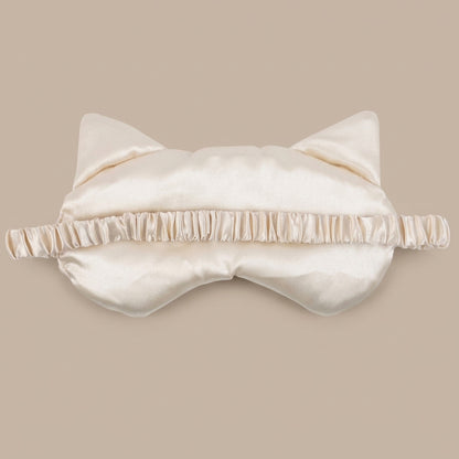 a back of an ivory satin sleep mask with cat ears and a ruffled elastic band, shown against a solid beige background.