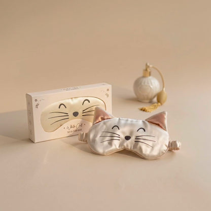 A satin cat-face sleep mask in a pale champagne color with embroidered black facial features and pink inner ears, shown in front of its white packaging box. A small decorative perfume bottle sits in the background on a warm beige surface.