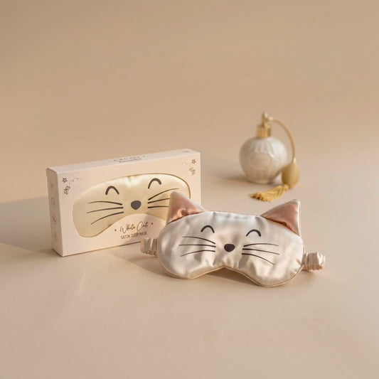 A satin cat-face sleep mask in a pale champagne color with embroidered black facial features and pink inner ears, shown in front of its white packaging box. A small decorative perfume bottle sits in the background on a warm beige surface.