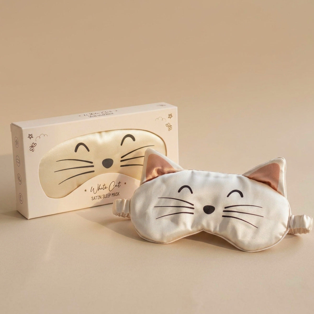 A white satin cat sleep mask with embroidered black facial features and pink inner ears, shown in front of its matching white packaging box on a warm beige background.