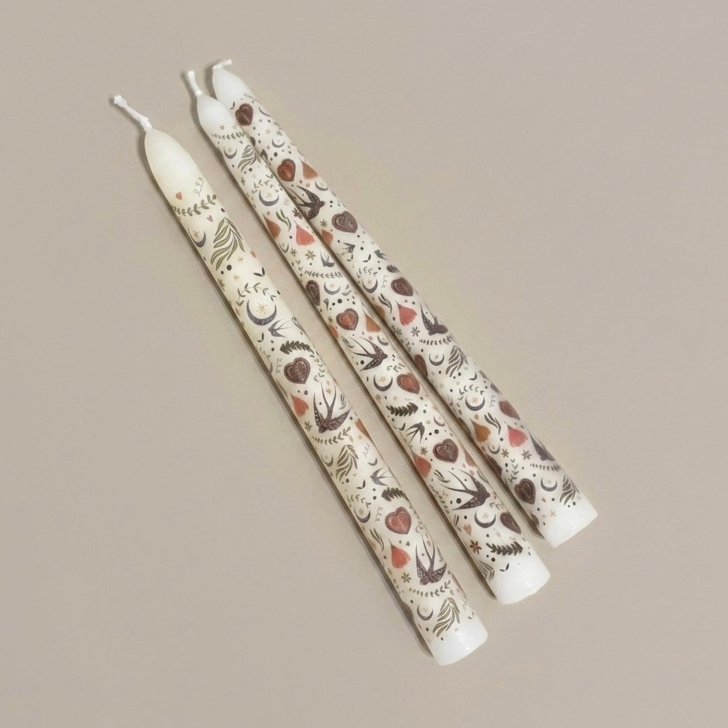Three cream candles with decorative bird and floral patterns on a beige background