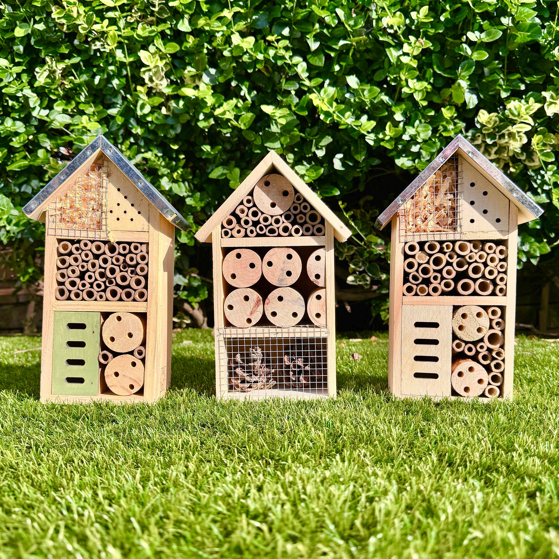 Three wooden bee hotels on grass with a green bush background