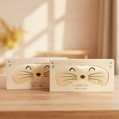Two white cat-themed sleep mask boxes on a wooden table, with a warm, softly blurred indoor background.