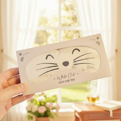 A hand holds a white sleep mask in its packaging, featuring a cute cat face design on the front. The background shows a softly blurred window with curtains, a small table, and flowers.
