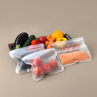 Four reusable silicone food storage bags with zipper closures, shown partially filled with groceries including tomatoes, carrots, peppers, and pasta. The bags are arranged on a plain beige background with a wooden board of fresh vegetables behind them.