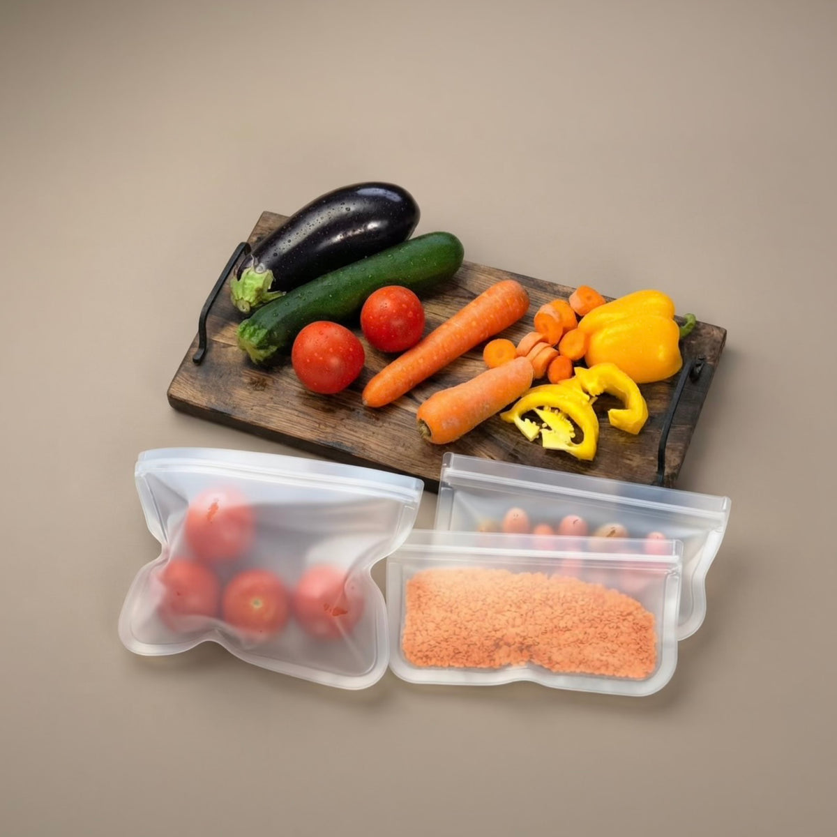Three reusable silicone storage bags filled with groceries, including tomatoes, carrots, a zucchini, an eggplant, and a yellow bell pepper, arranged on a beige background with a wooden cutting board in the background.