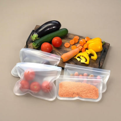 Four reusable silicone zip-top storage bags filled with tomatoes, sliced carrots, yellow bell pepper, and orange powder, arranged on a beige surface with a wooden cutting board holding fresh vegetables in the background.